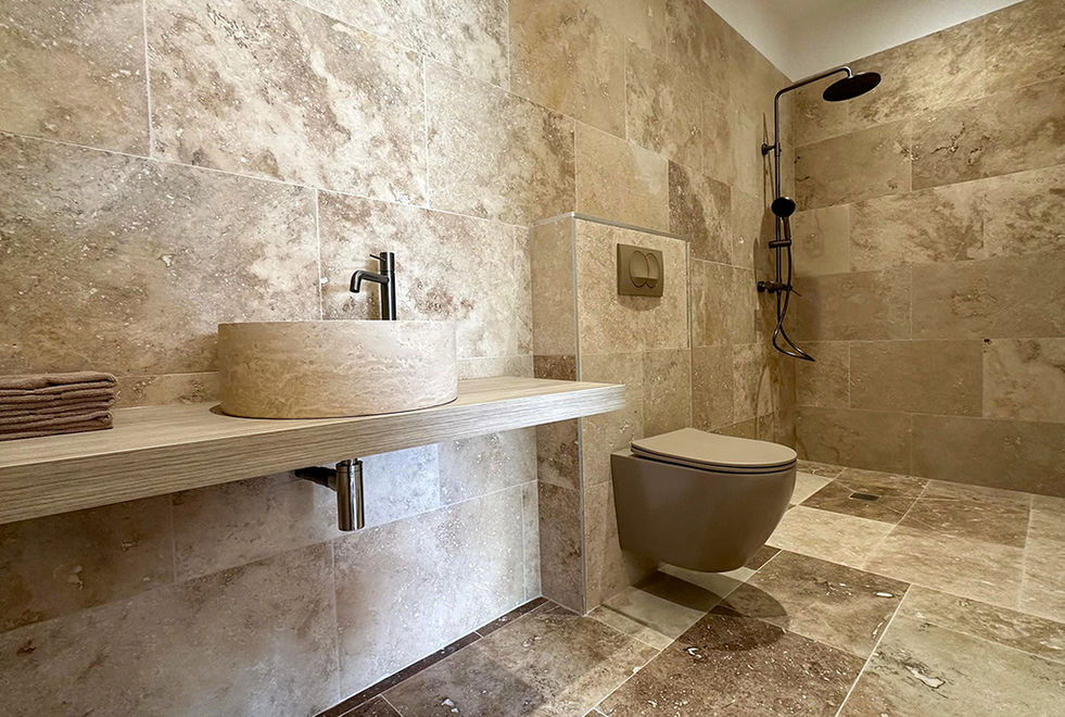 Salle de bain en pierre travertin avec vasque ronde, WC suspendu taupe et colonne de douche noire mat, villa de luxe Solenzara Corse