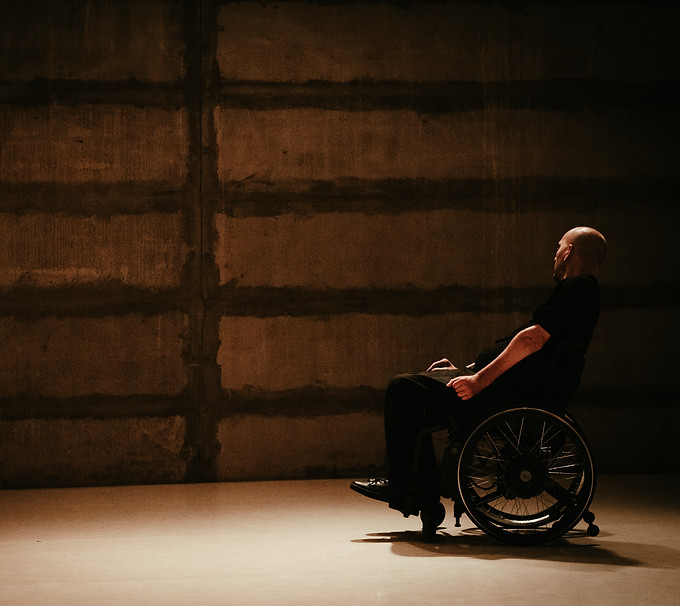A dancer using a wheelchair is seen in profile moving to the left, along a brick wall.