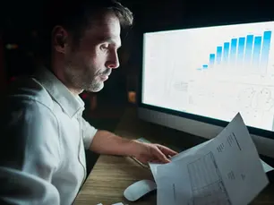 Homem de perfil, usando camisa branca, analisa documentos impressos à noite em uma mesa de escritório, enquanto observa um monitor grande com gráficos e dados financeiros iluminados.