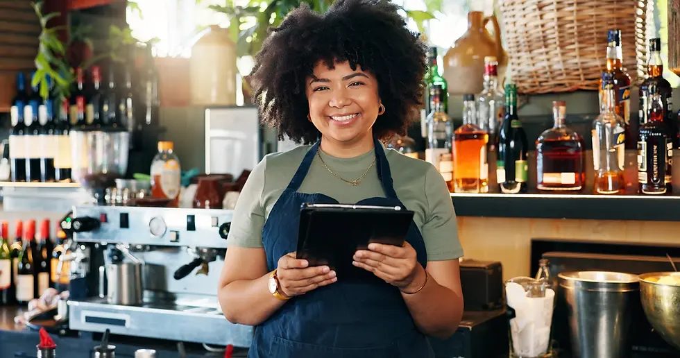 Mulher sorrindo atrás do balcão de um bar ou cafeteria, usando avental e segurando um tablet para controle de pedidos ou estoque, com garrafas de bebidas e máquina de café ao fundo.