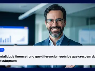 Homem de terno azul sorrindo em um escritório, com colegas ao fundo e gráficos de crescimento projetados na tela.