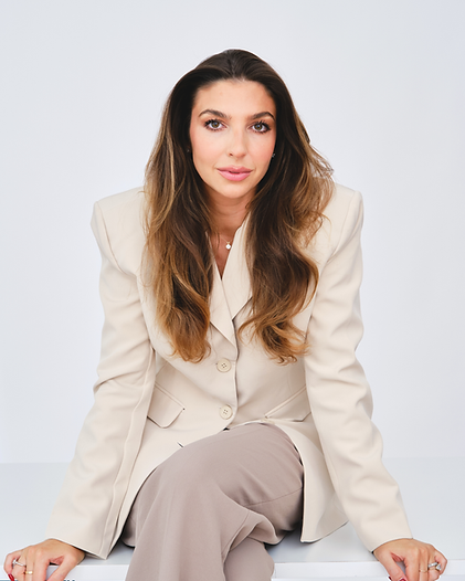 A woman with long brown hair sits on a white surface, wearing a light beige blazer and pants, against a white background.