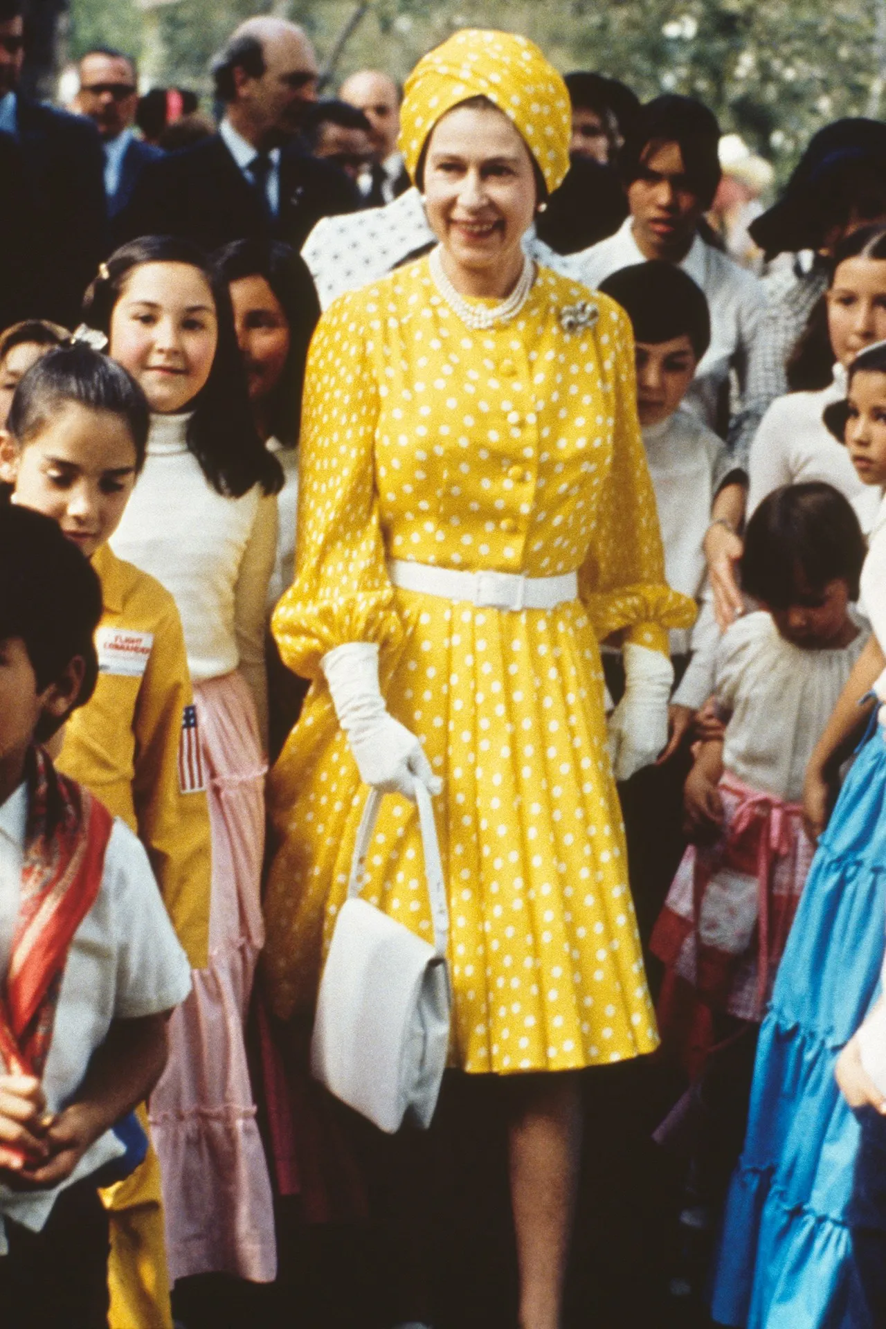 The Queen wearing a yellow polka dot dress and turban, with children on her visit to Mexico in 1975