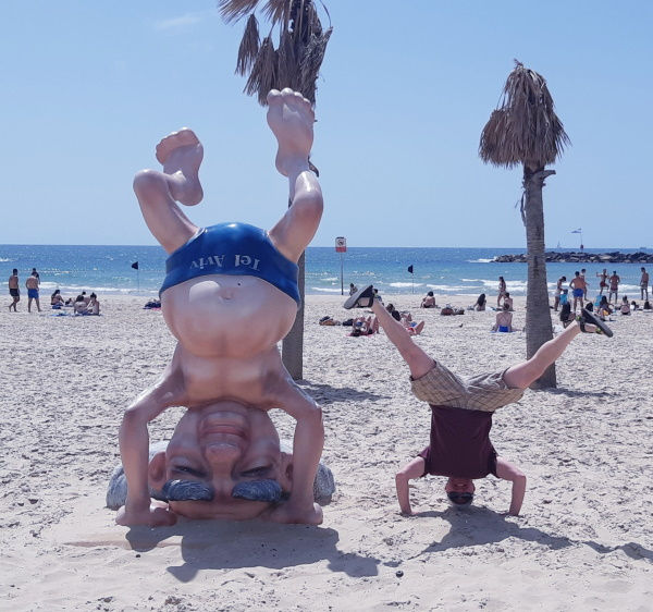 A young man performing a headstand on the sand next to a sculpture of Ben Gurion on his head