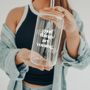 May include: A clear glass can-shaped tumbler with a wooden lid and a straw. The tumbler is filled with a light brown liquid. White text on the front of the glass reads 'good things are coming' with flower graphics. The glass is held by a person with a bracelet.