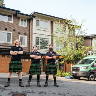 Three smiling workers wearing tartan kilts and black shirts stand confidently with arms crossed in front of residential homes, with a branded work van in the background