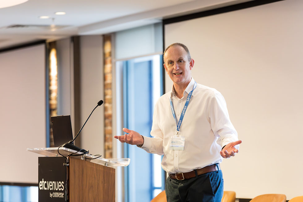 A man speaks at a podium labeled wearing a white shirt and lanyard. Bright conference room with a large window.