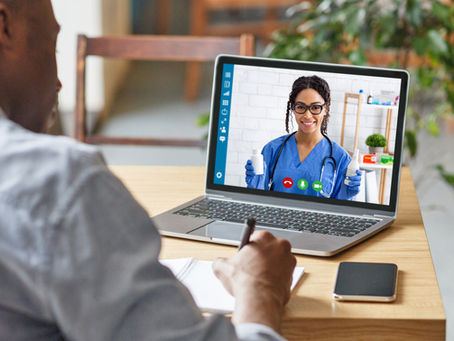 Man taking notes during a video call with a smiling female doctor on a laptop. Doctor in blue scrubs holds a bottle, office background.