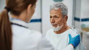 A doctor examines an elderly man's shoulder during a medical consultation in a clinical setting.