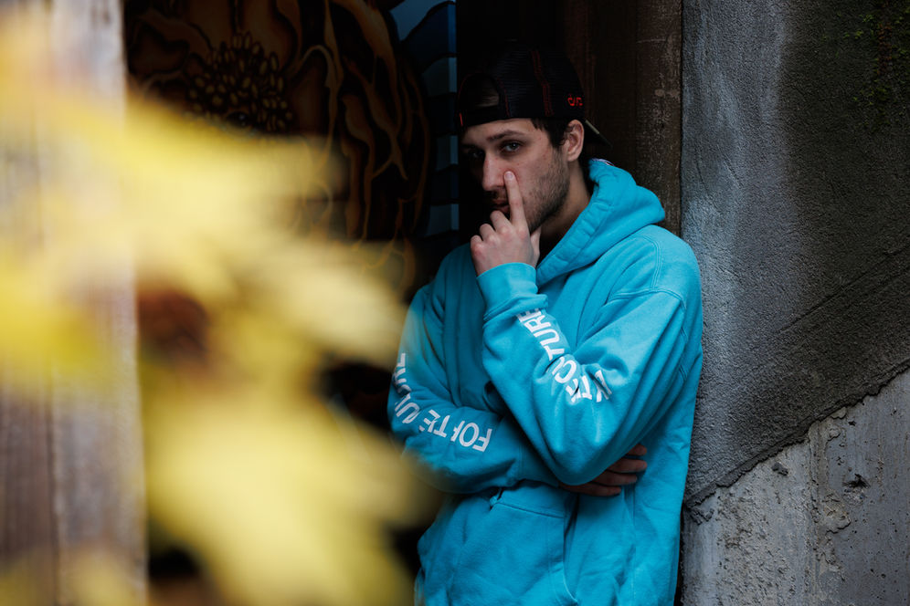 Young man in blue hoodie with text, leaning against a wall, in a thought.