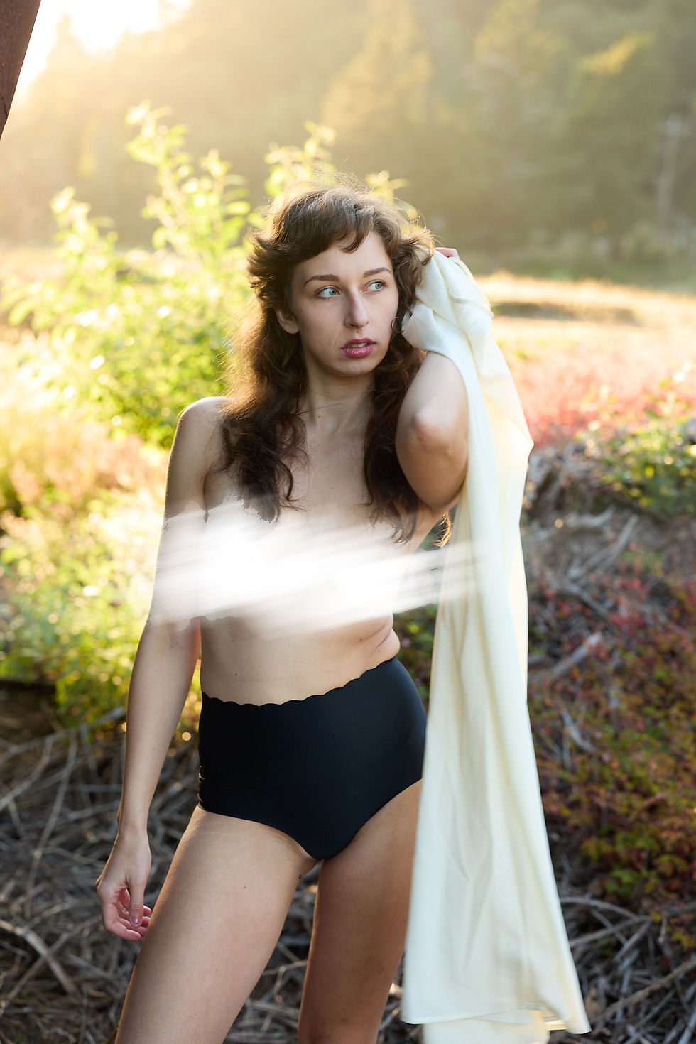 Woman in black high-waisted swimsuit bottoms holds white fabric outdoors in warm sunlight, looking off thoughtfully.