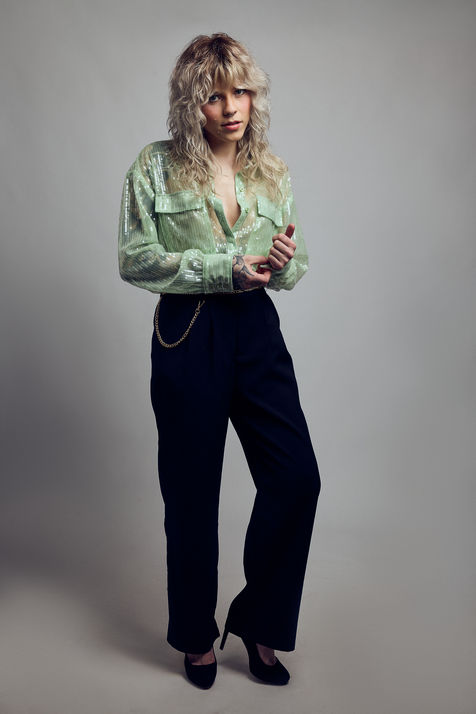 Woman wearing a green shirt, black pants, and heels, FASHION, standing against gray backdrop.