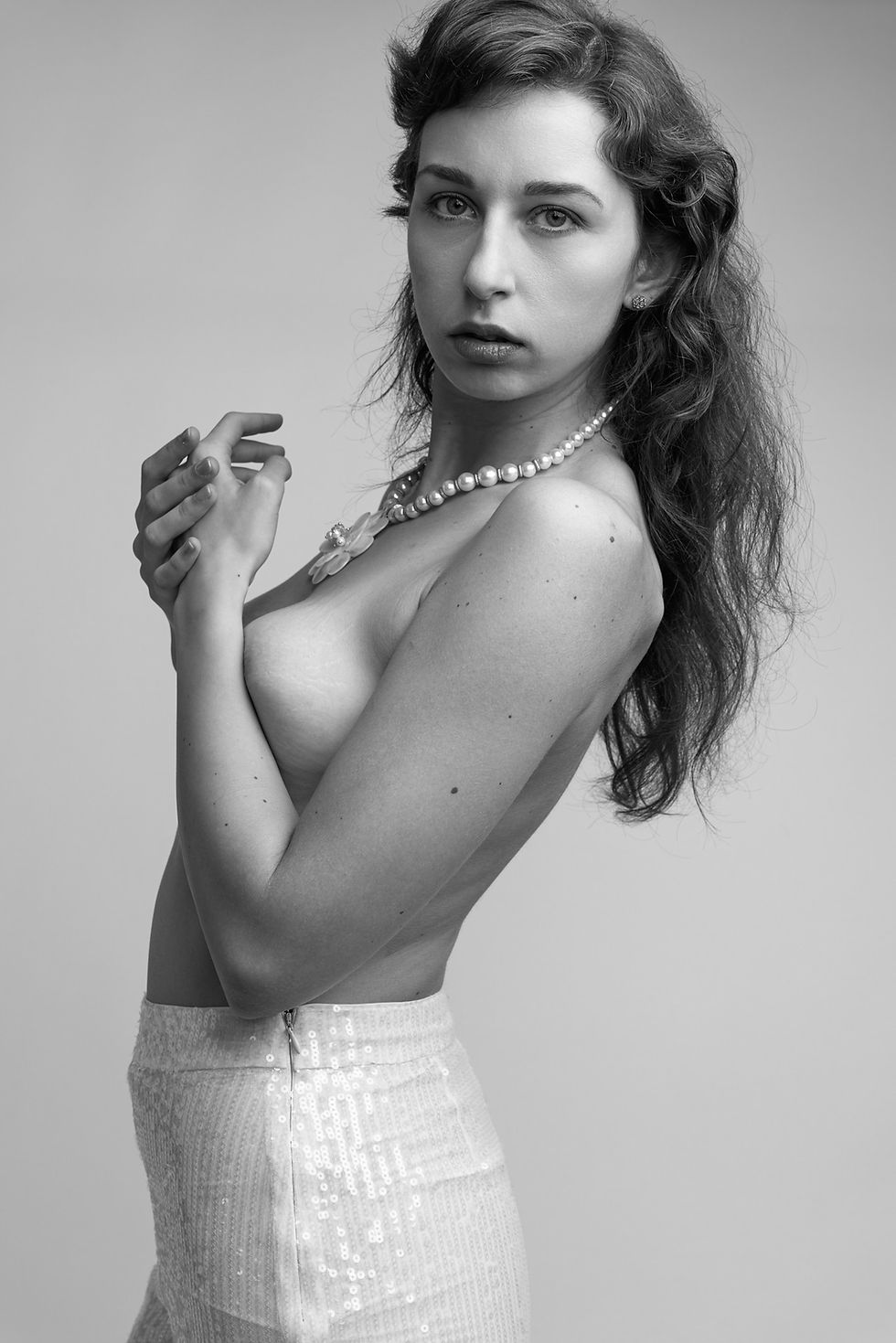 Black-and-white studio portrait of a woman in pearls and a sequined skirt, arms crossed, gazing seriously at the camera.