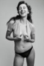 Black-and-white studio portrait of a laughing woman in an open knit top and black underwear against a plain gray background.
