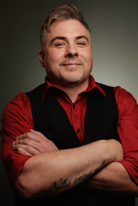 Man smiles with arms crossed, wearing a red shirt and vest. Nick Johnson Photo