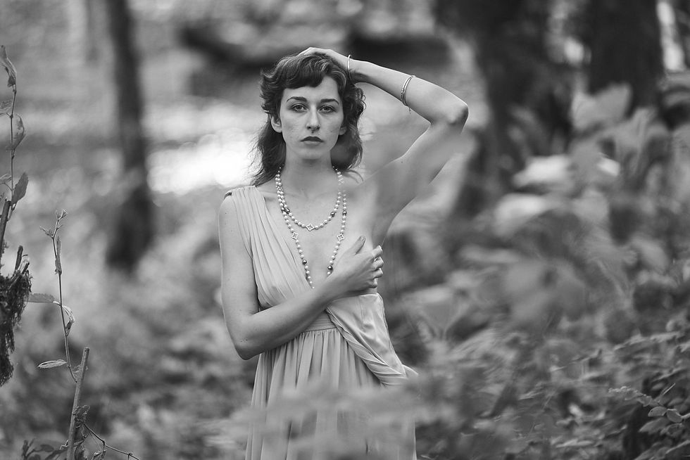 Black and white outdoor portrait of Lindsey standing among ferns in a flowing dress and pearl necklace.