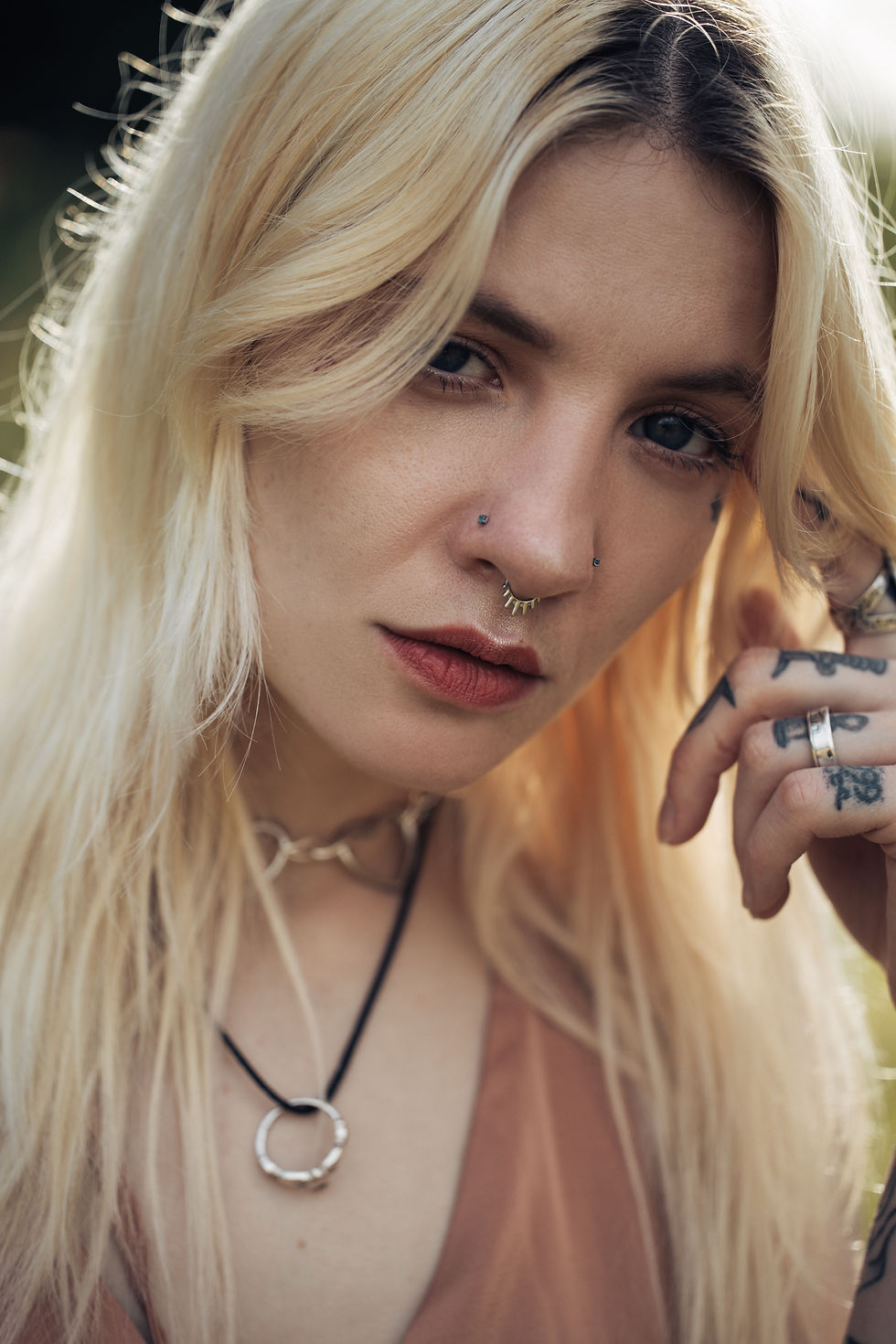 Blonde woman with nose piercings and tattoos gazes intently. She's outdoors, wearing a brown top and necklace, with a soft background.