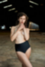 Portrait of a woman in a dim warehouse, hands clasped at her chin, wearing black high-waisted bottoms and gazing seriously.