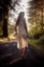Barefoot woman in a white dress looks back on a sunlit forest road, with warm golden light and blurred trees.