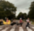 Four golfers cross a zebra crossing on a cloudy, tree-lined road, carrying golf bags; one wears a bright yellow inflatable float.