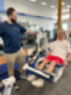 Man in a blue shirt assists another man in red shorts using a leg extension machine in a gym. Bright, well-lit interior setting.