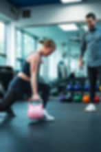 Woman doing a kettlebell lunge in a gym, observed by a trainer. Bright setting with colorful kettlebells in the background.