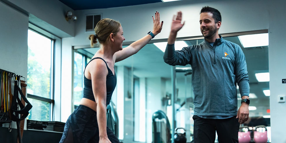 Man and woman high-five in a gym, smiling. She wears a sports bra, he wears a gray long-sleeve. Bright, modern setting with exercise gear.