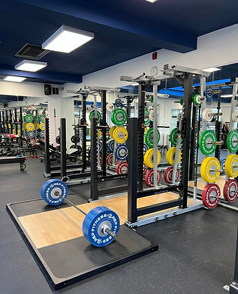 a well stocked gym with colorful bumber plates lining 3 squat racks. A deadlift platform is loaded with a barbell.
