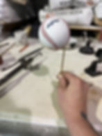 A white hand holds a brass mount that is holding a baseball. There are various workshop supplies in the background. 