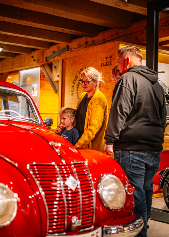 Familie betrachtet in einer Holzhalle einen roten Oldtimer; Mädchen, Frau und Mann stehen nah am Wagen.
