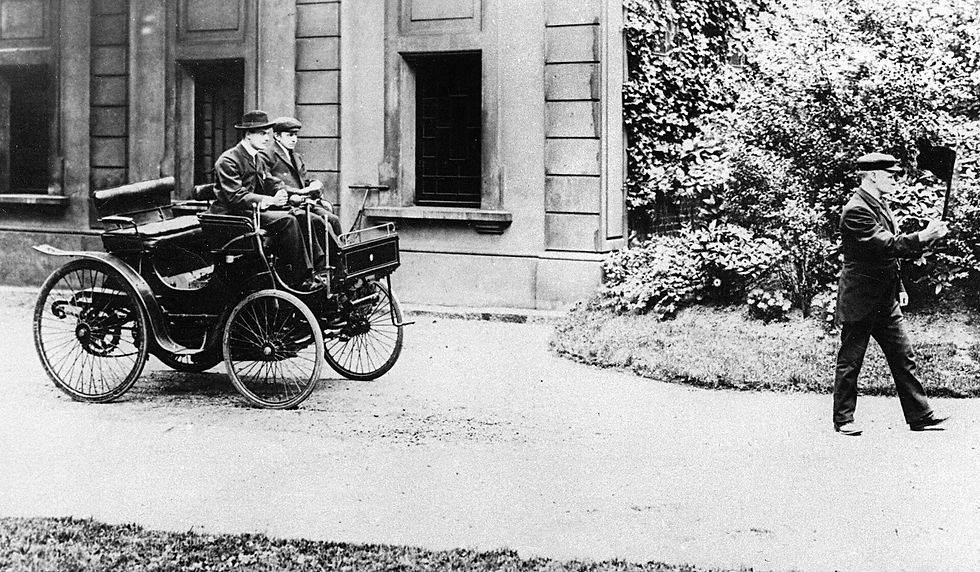 Historische Schwarz-Weiß-Aufnahme eines frühen Automobils vor einem Gebäude, besetzt mit zwei Personen, während rechts eine weitere Person über den Weg geht.