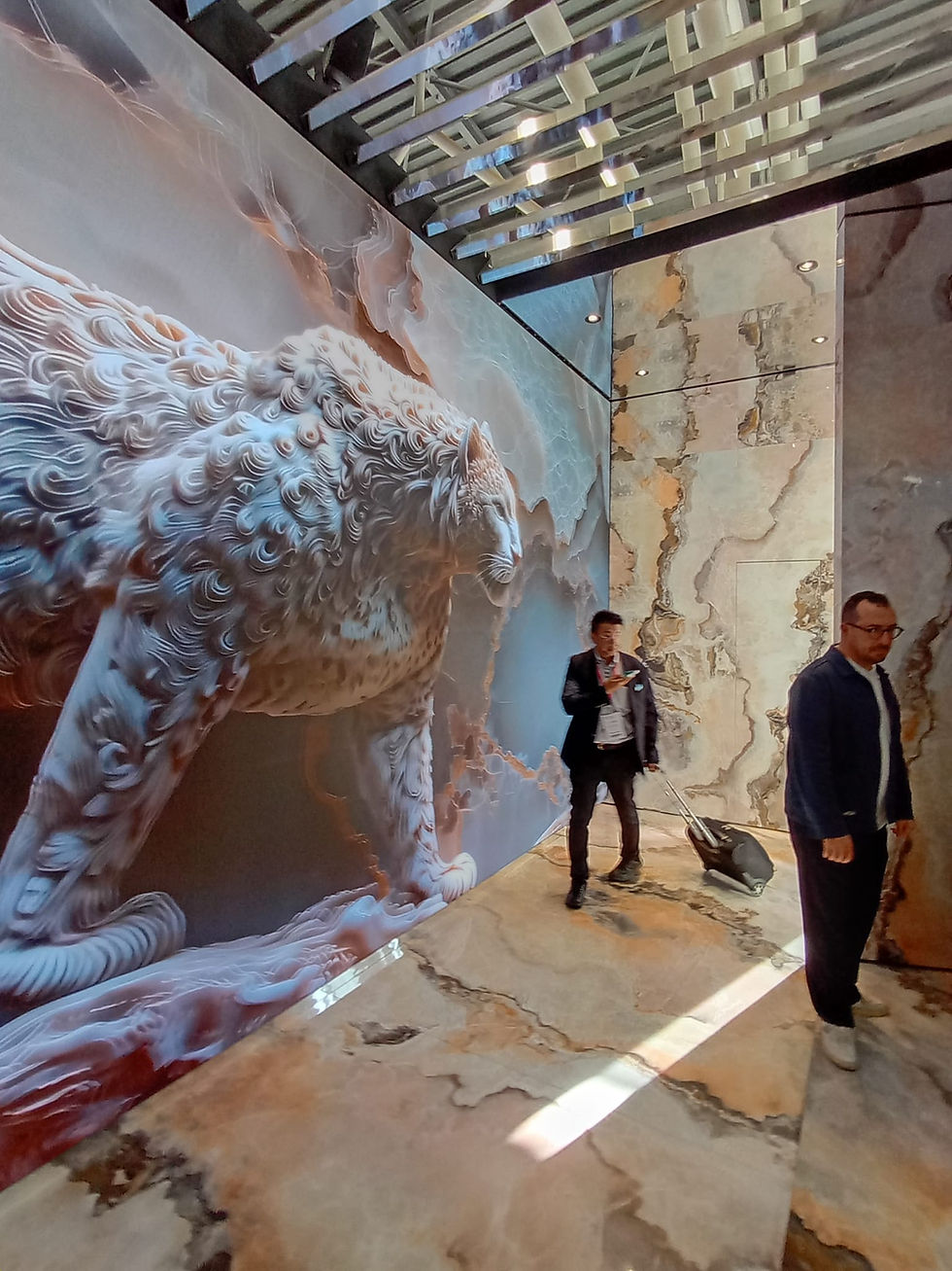 A modern art installation featuring a detailed, swirling depiction of a mythological creature on a wall, with individuals interacting in the brightly lit, marble-floored space beneath a geometric ceiling.