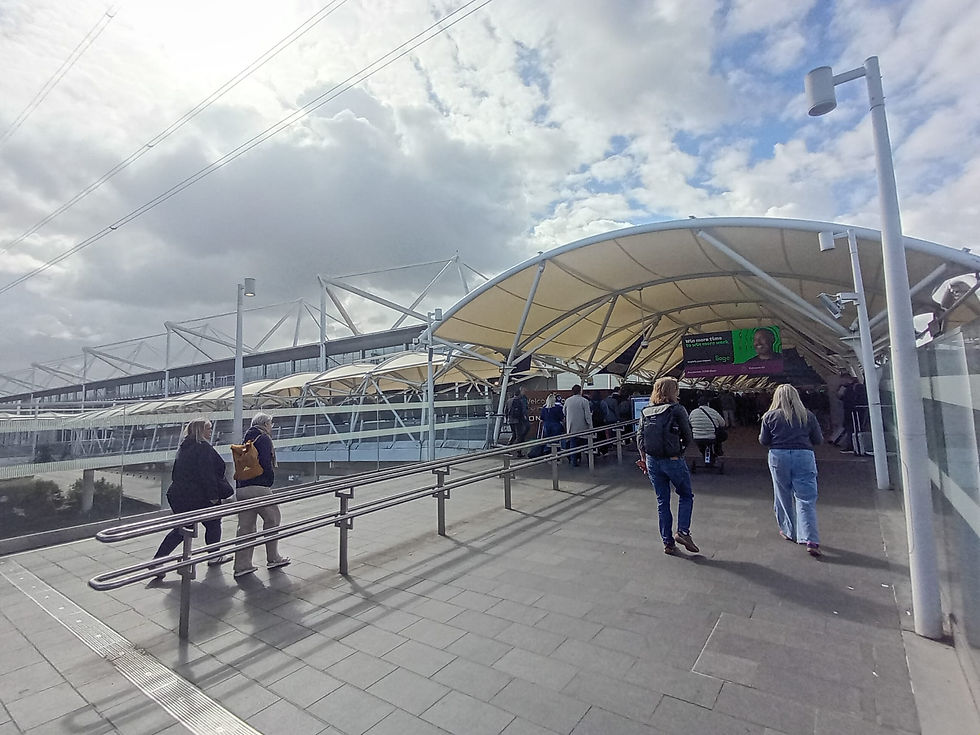 Under a cloudy sky, travelers navigate a bustling transport hub, moving towards the ExCeL London exhibition centre entrance