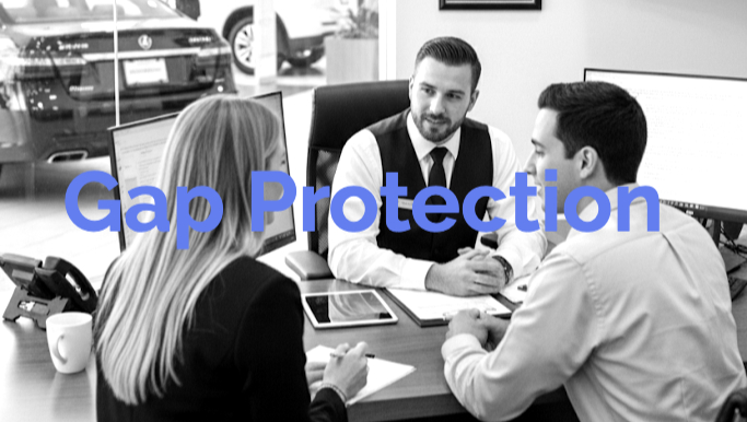 Salesman meeting with couple at desk in car dealership showroom, vehicles visible in background.
