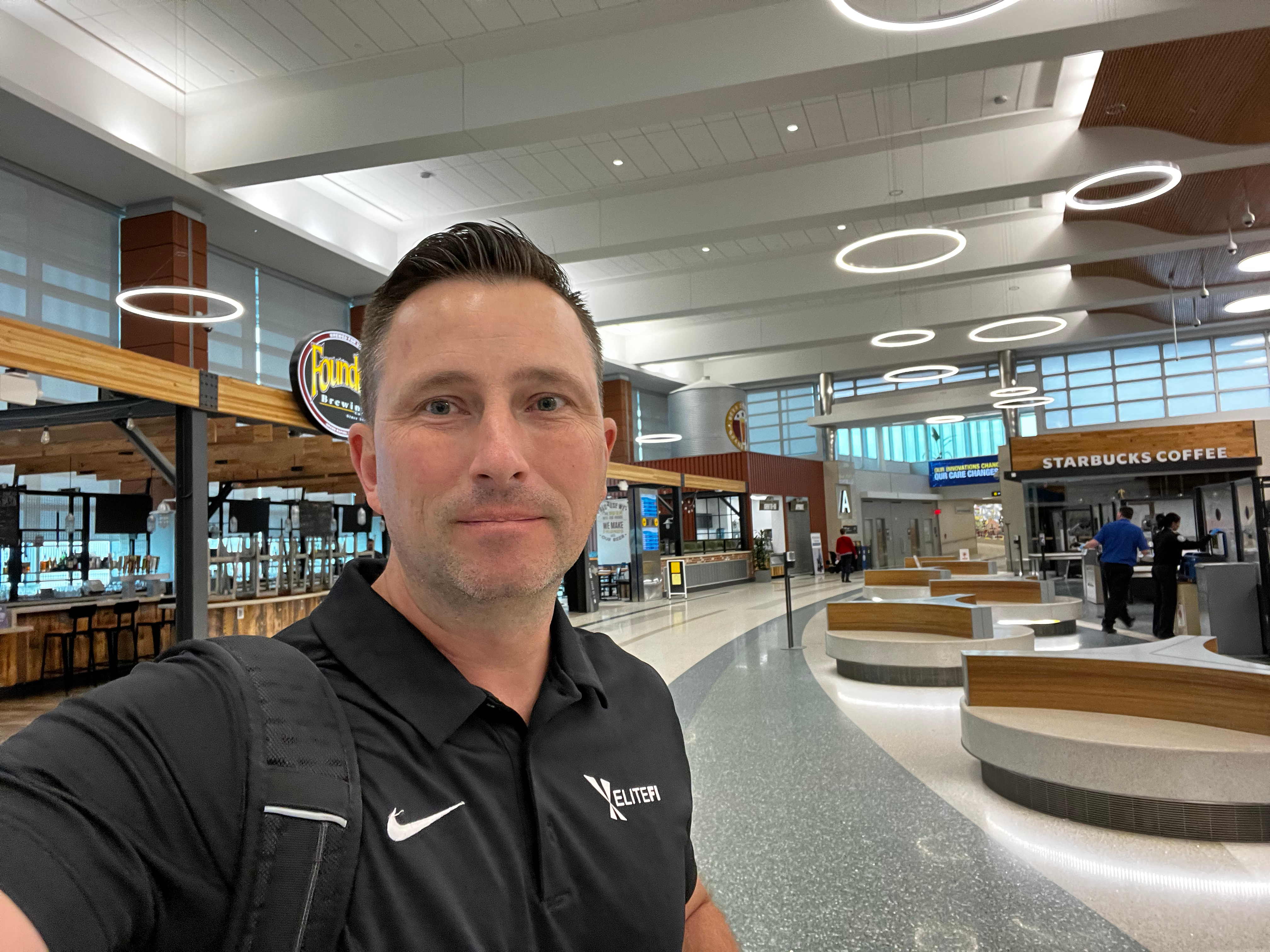 Man in black Nike polo taking selfie in modern airport terminal with Starbucks, restaurants, and circular seating areas.