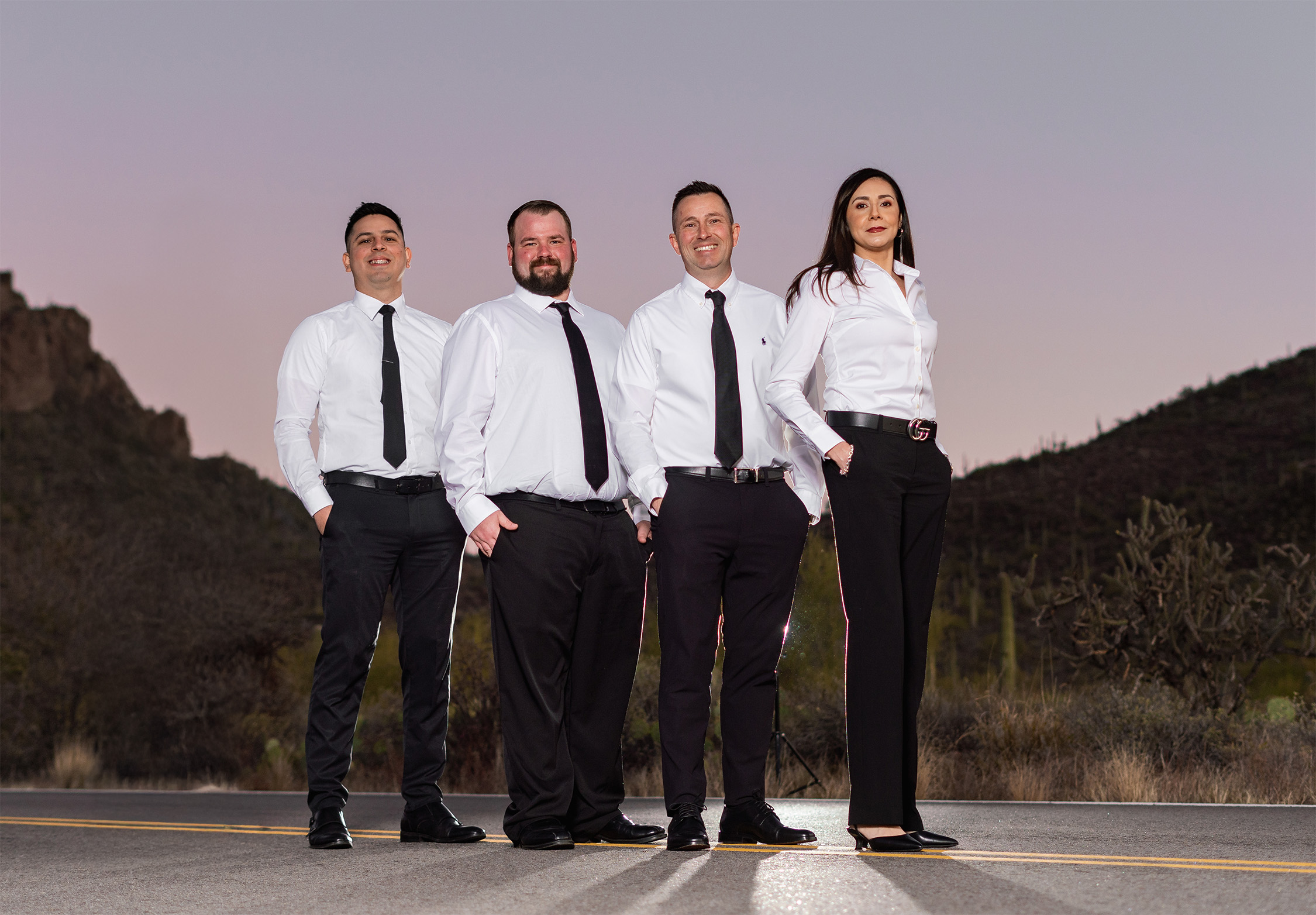 Four business professionals in white shirts and dark ties standing on a desert highway with mountains in background at dusk.