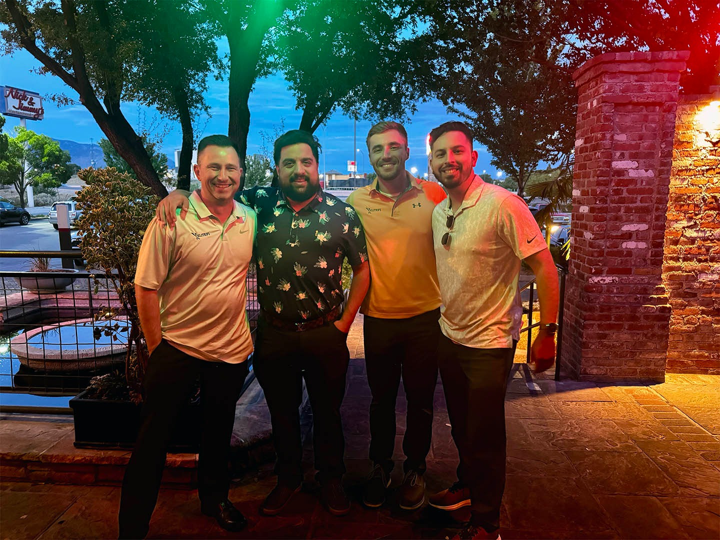 Four men standing together outdoors at dusk, smiling at camera, with brick building, large trees, and twilight sky in background.