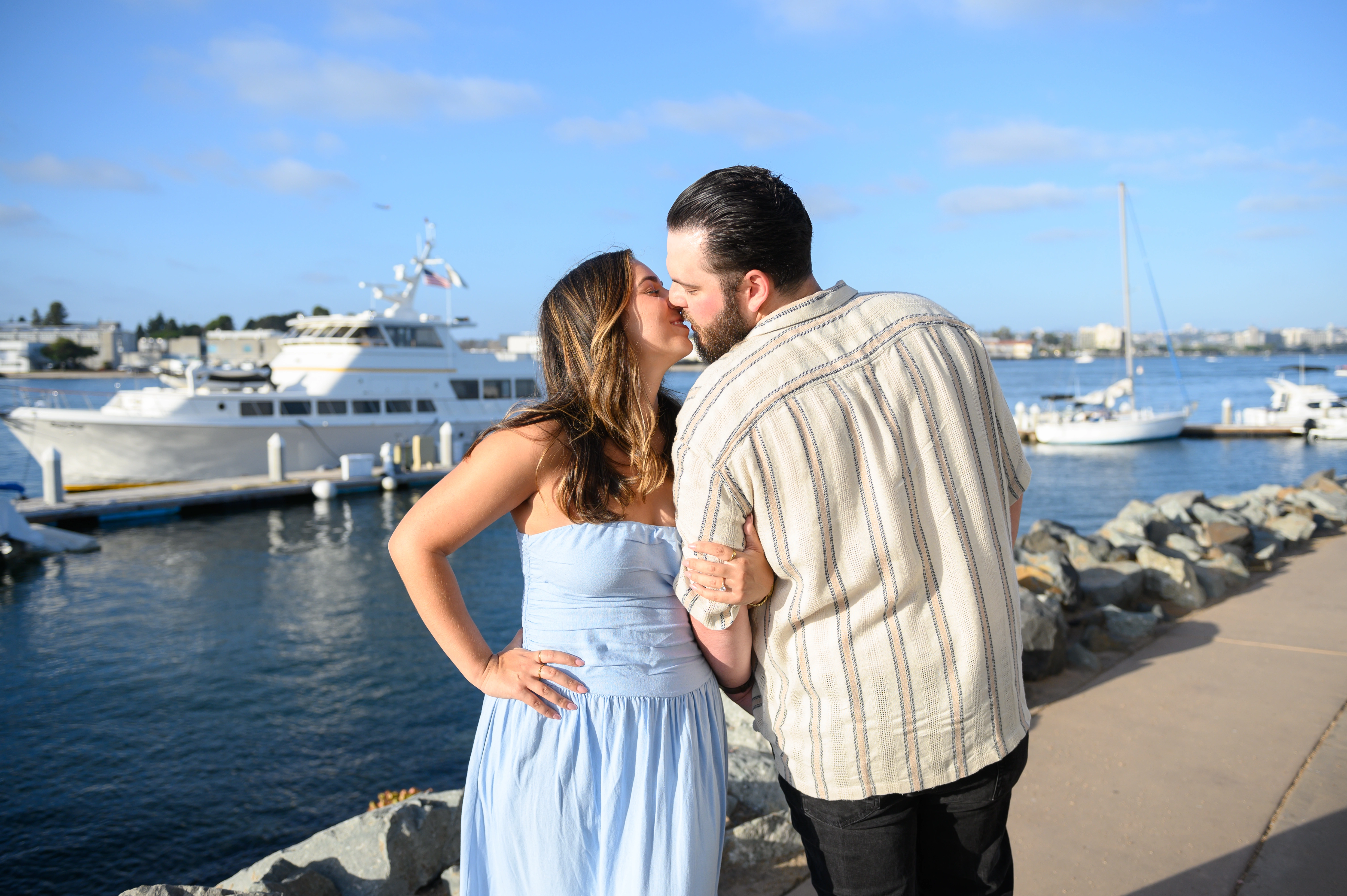 Engaged couple sharing a kiss while holding hands by the water in San Diego, photographed by an affordable San Diego proposal photographer.