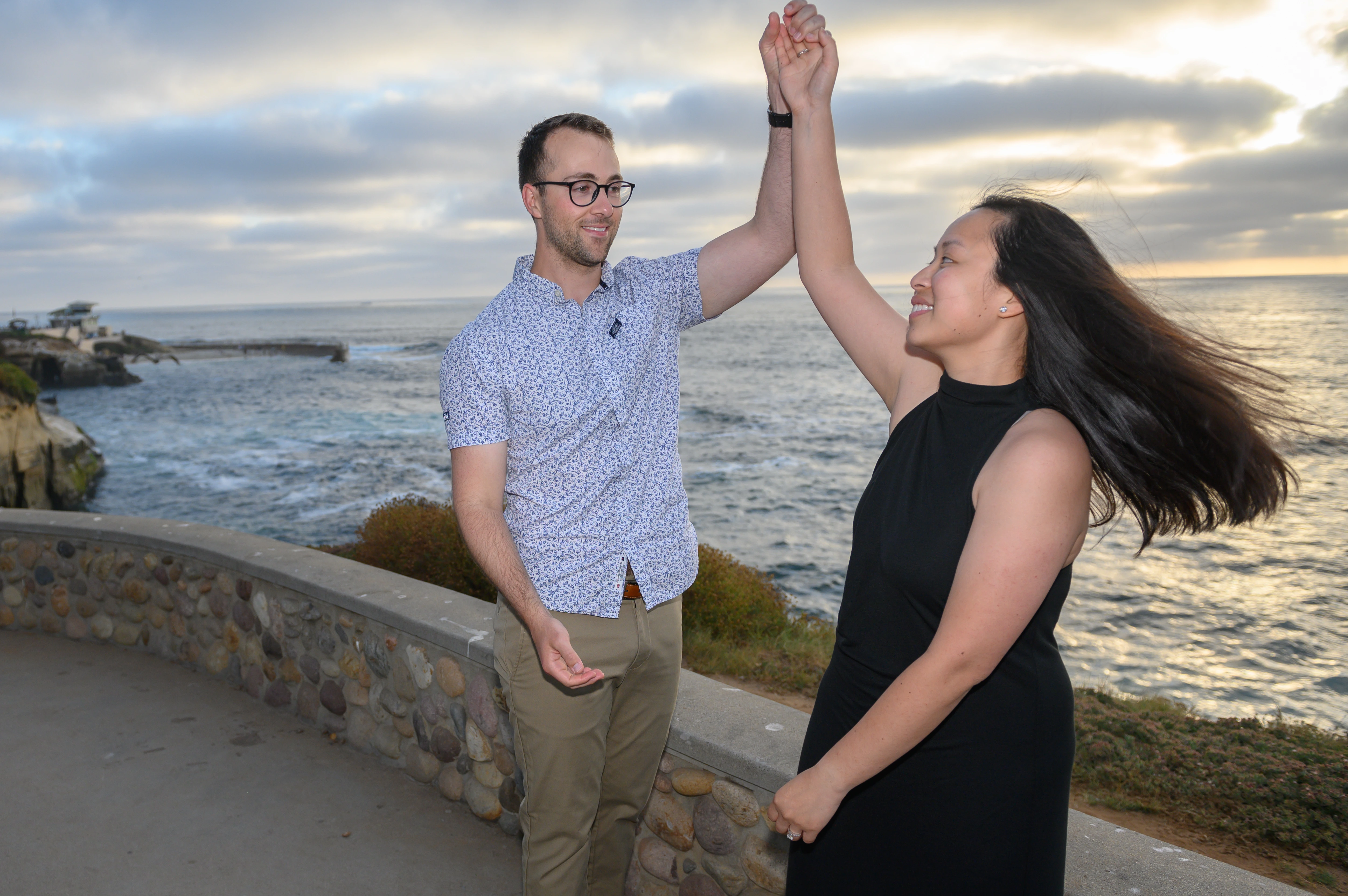 The couple joyfully dances together by the ocean, with the waves and a cloudy sky in the background