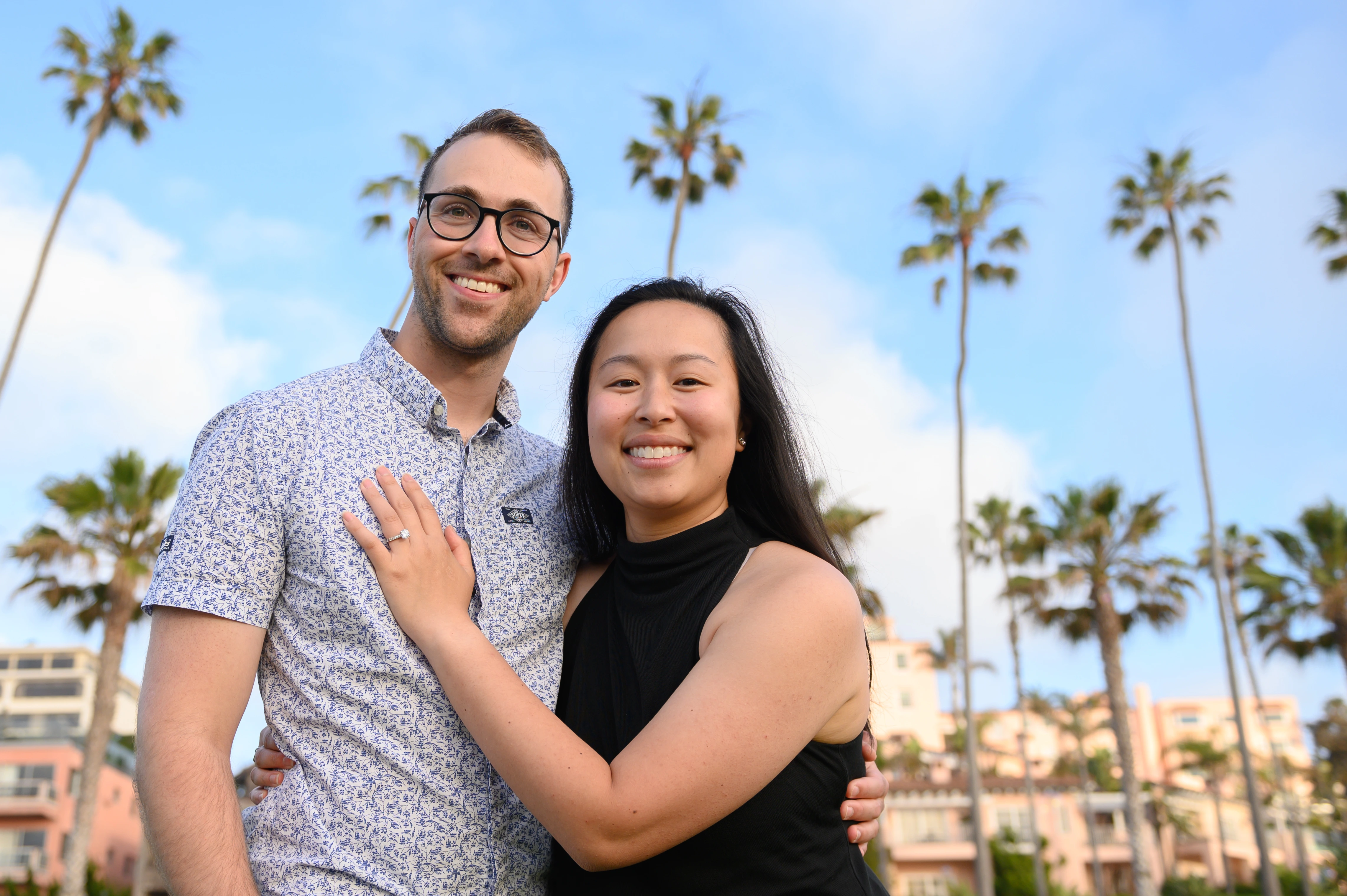 The couple stands close together, smiling against a backdrop of palm trees and a colorful building