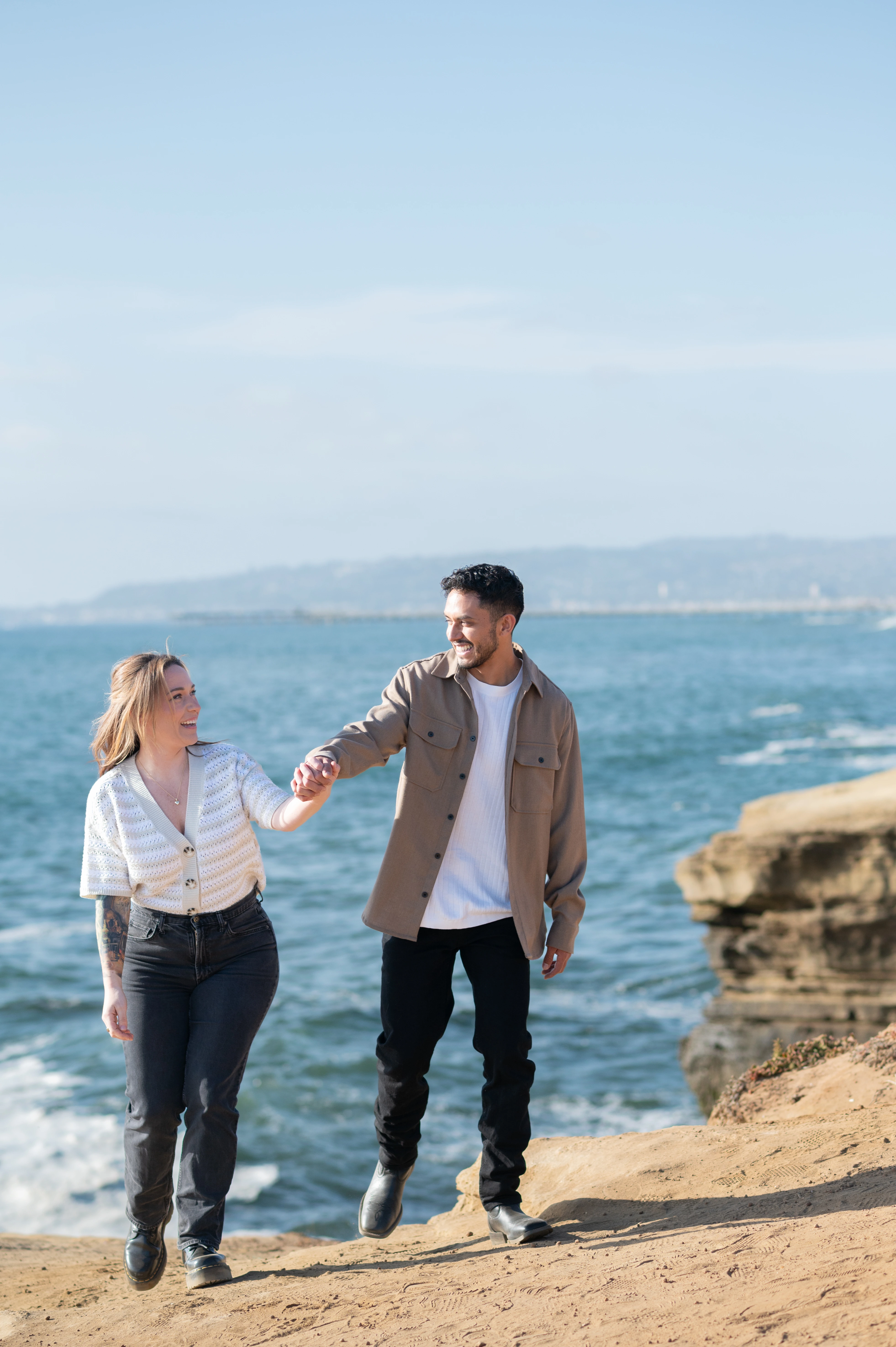 A couple walks along the rugged coastline of Sunset Cliffs, holding hands and sharing laughter, embodying the joy and romance of their engagement journey in San Diego.