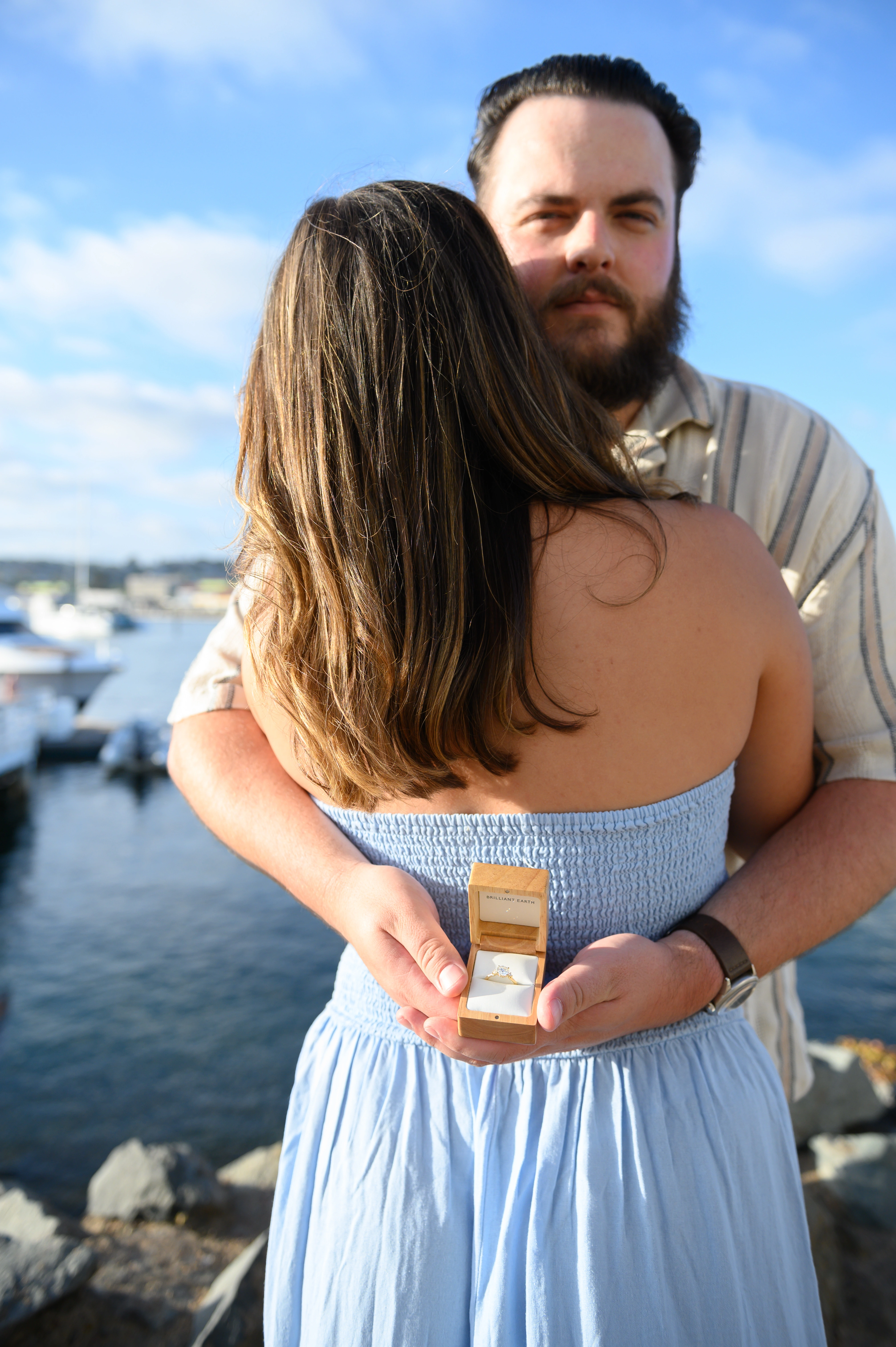 Man holding an engagement ring while embracing his girlfriend at a scenic San Diego waterfront, captured by the best San Diego proposal photographer.