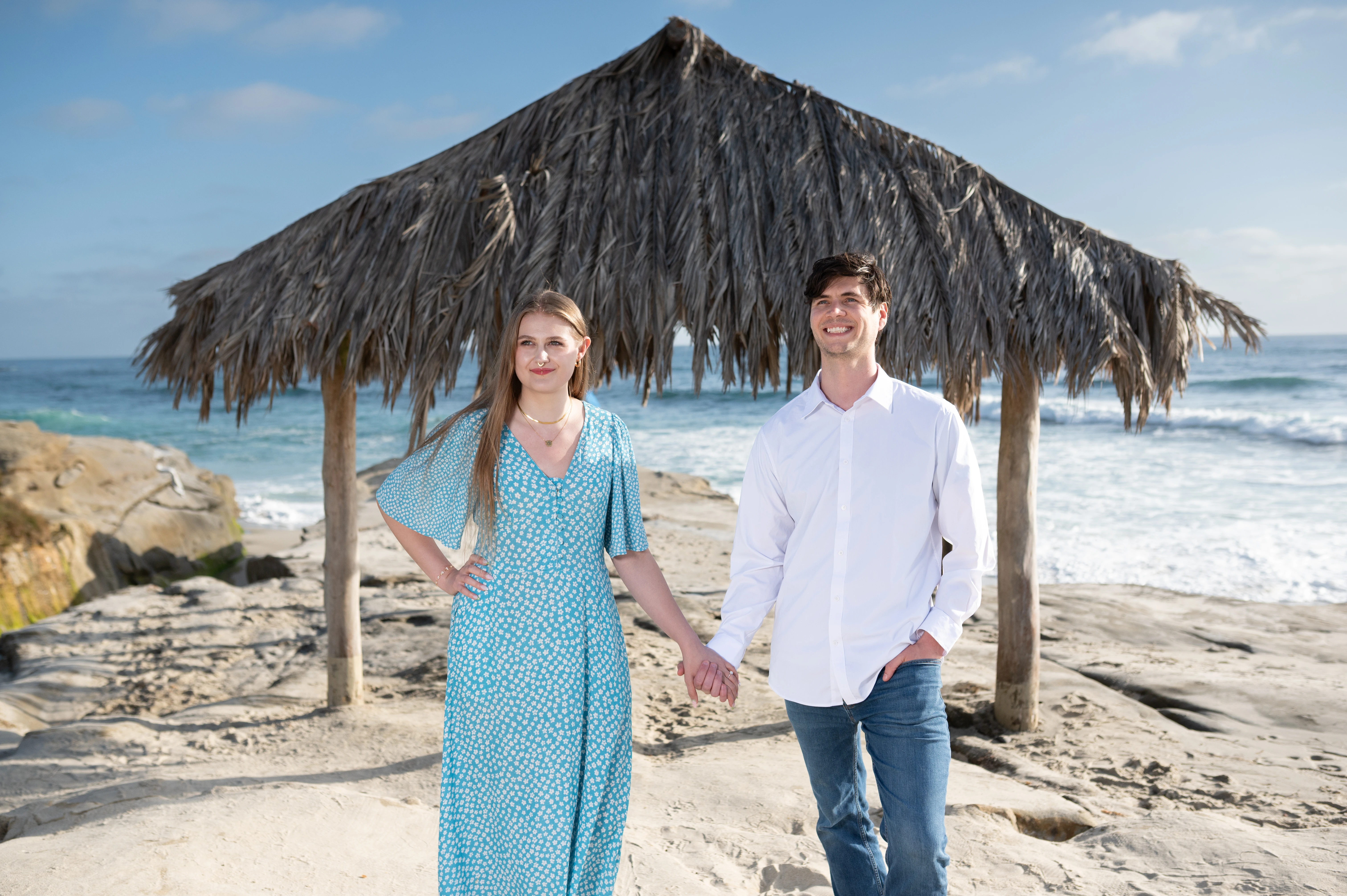 A couple stands hand in hand under a thatched roof structure by the beach, with waves crashing in the background