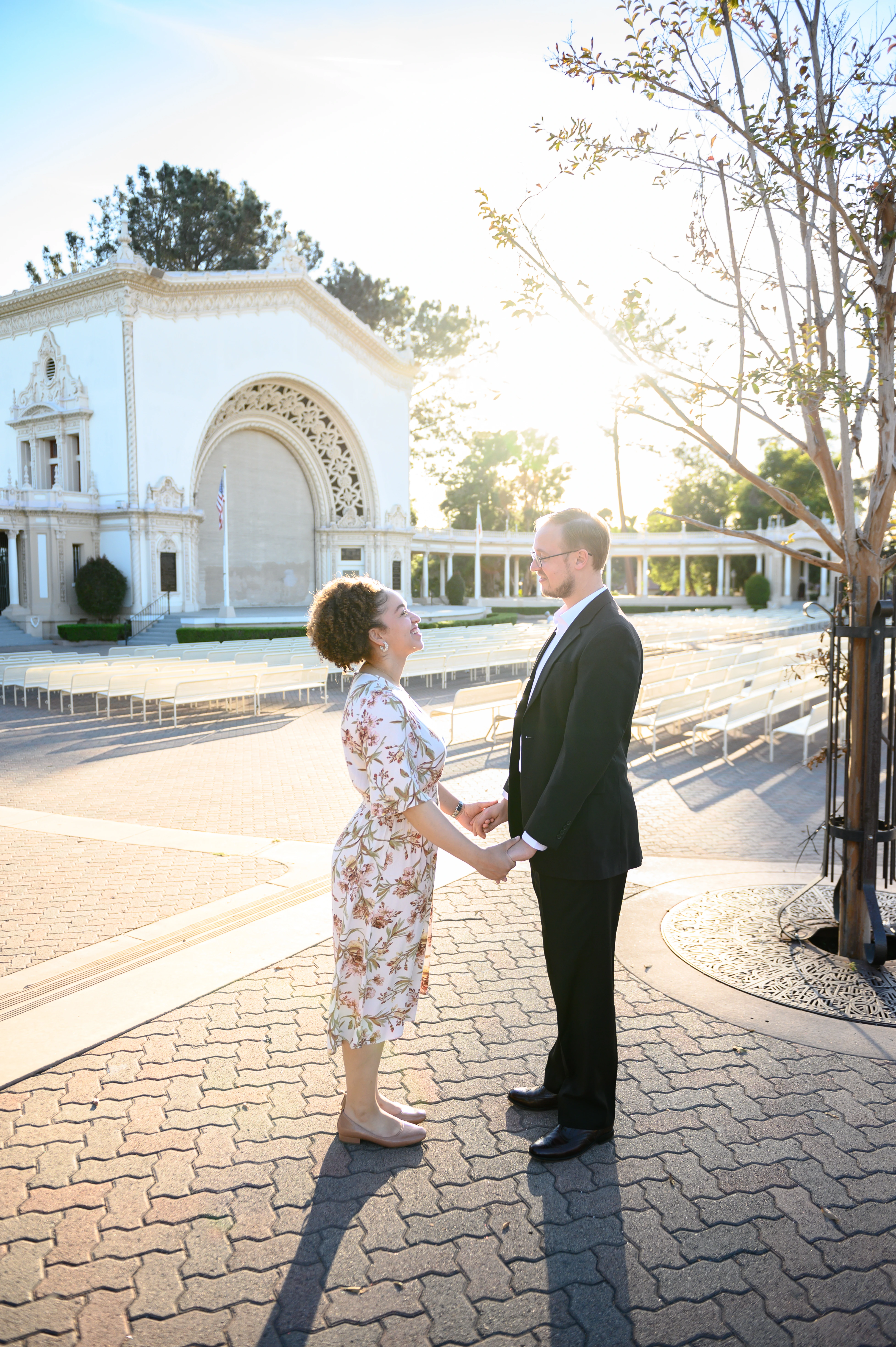 The couple holds hands and gazes at each other in a sunlit outdoor setting, with a beautiful architectural backdrop in the distance.
