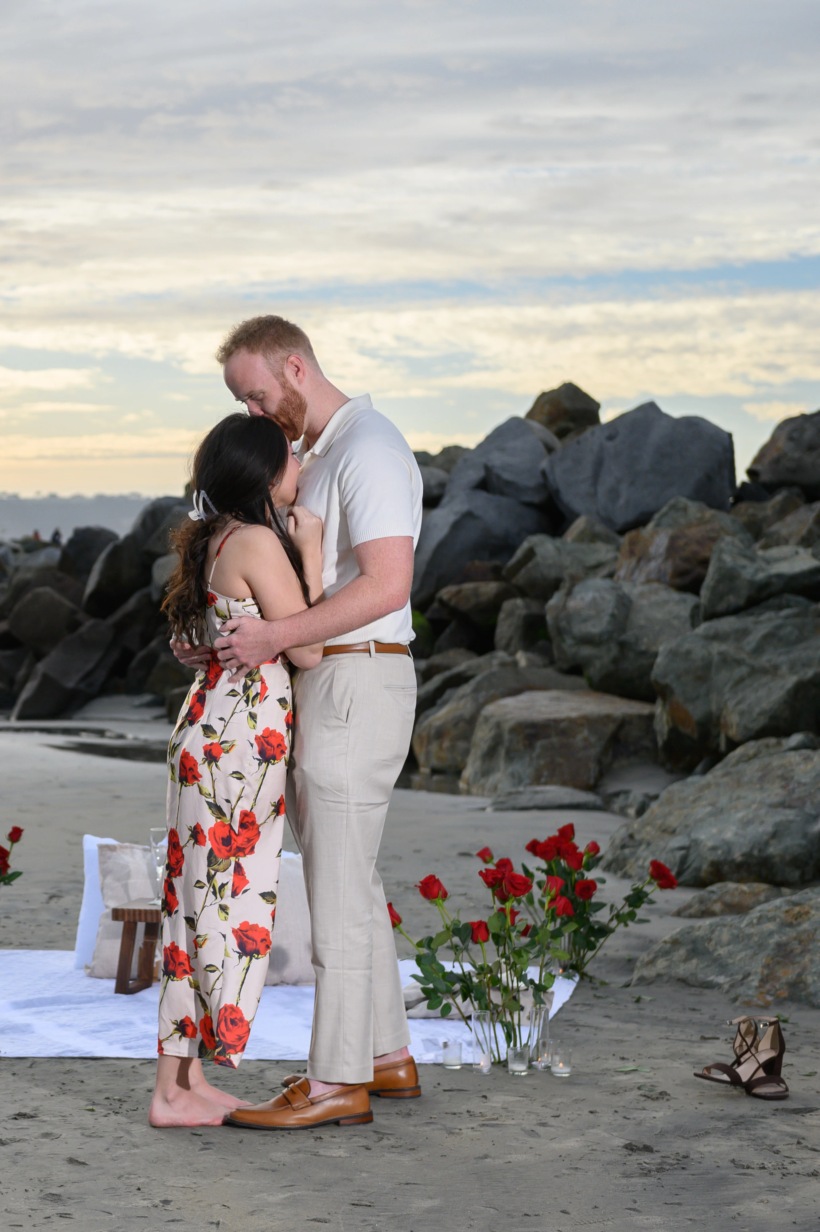 A couple embraces on the beach surrounded by roses, with rocky formations in the background and a colorful sky at sunset