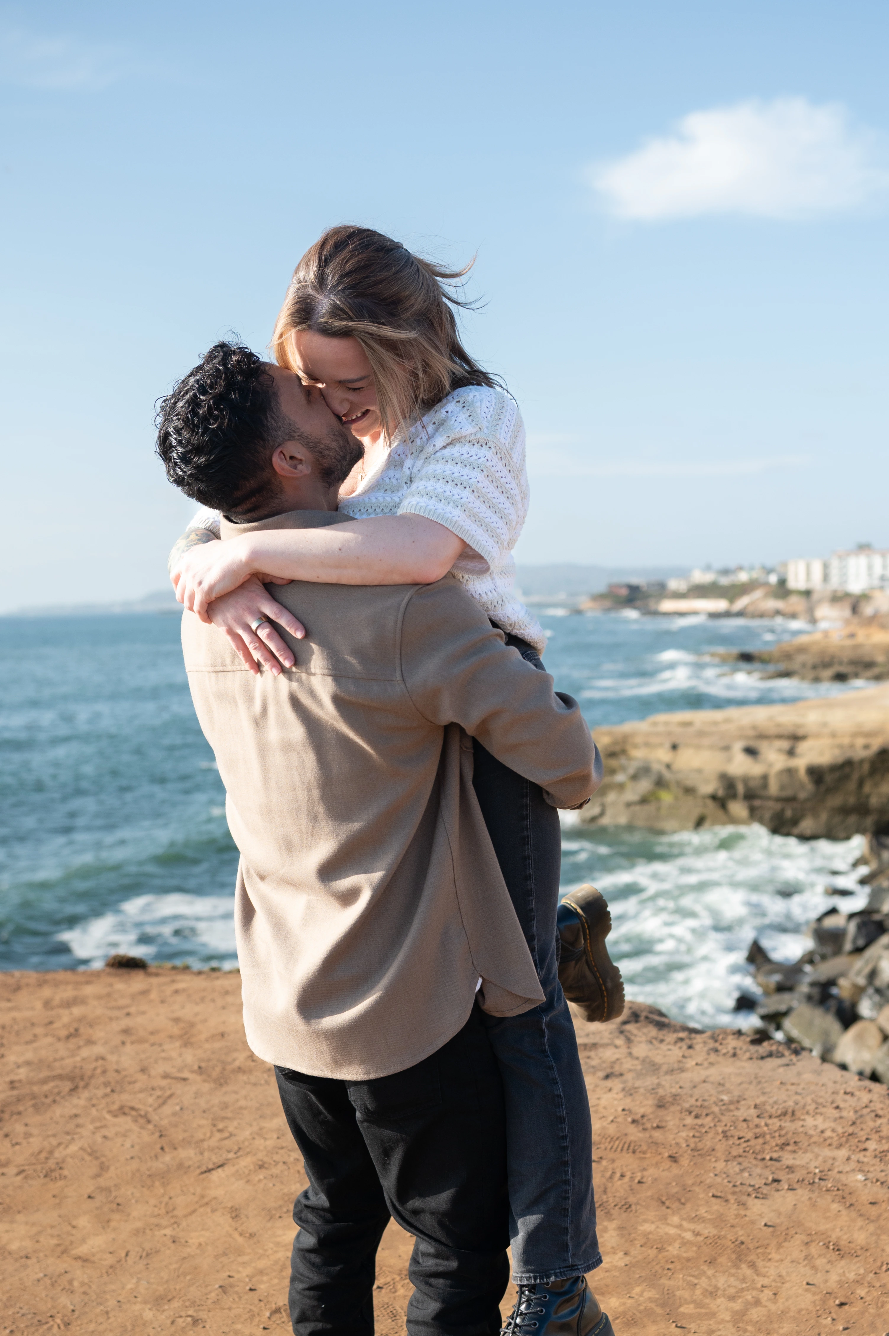 In a joyful embrace, a man lifts his partner off the ground against the stunning backdrop of the ocean at Sunset Cliffs, showcasing the love and excitement of their engagement.