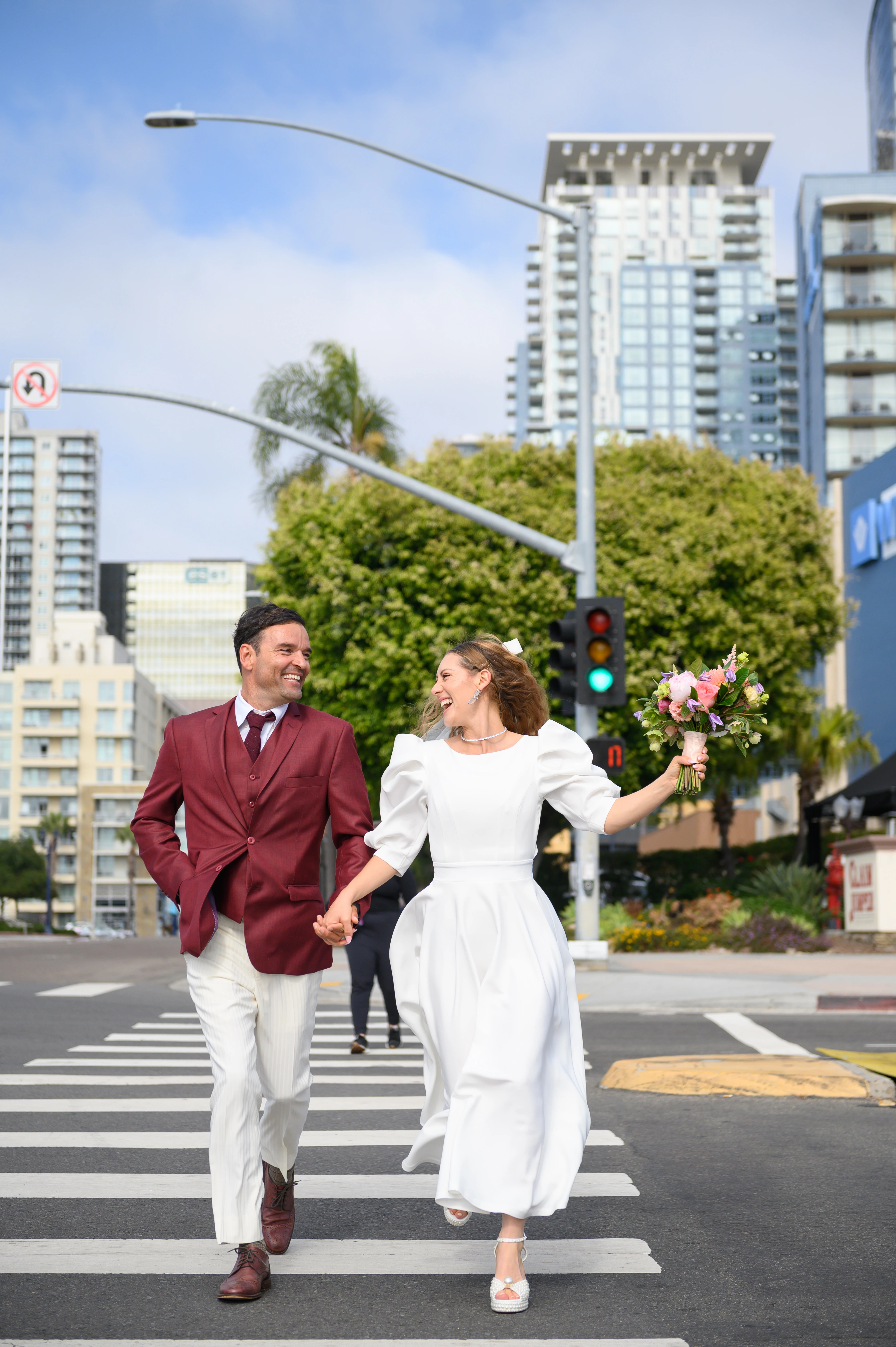 A joyful couple skipping hand-in-hand across a pedestrian crossing in downtown San Diego, with city buildings and street signs in the background
