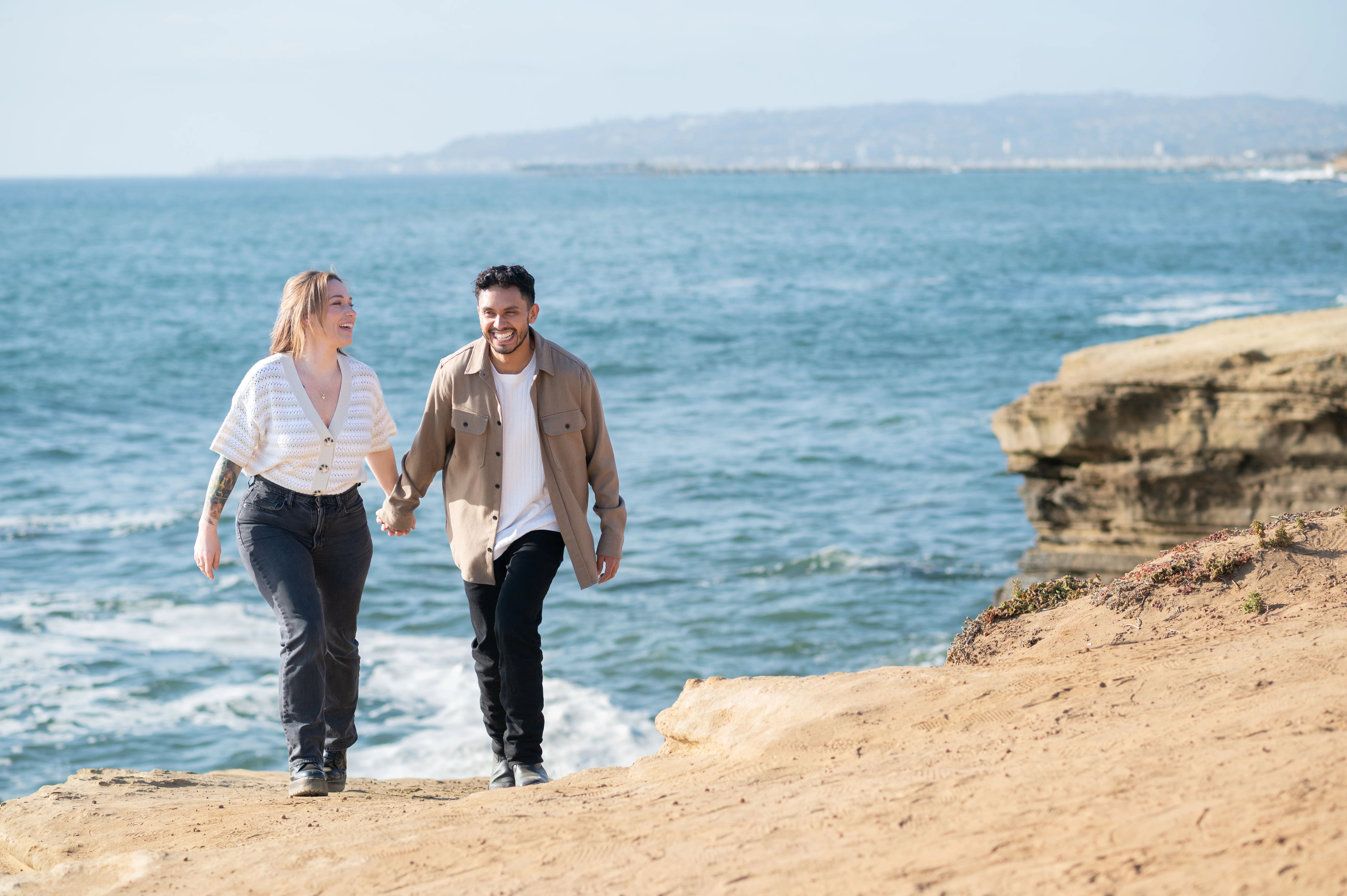 A joyful couple walks hand-in-hand along the sandy cliffs of Sunset Cliffs, with the sparkling ocean in the background. Their laughter and connection reflect the excitement of their engagement