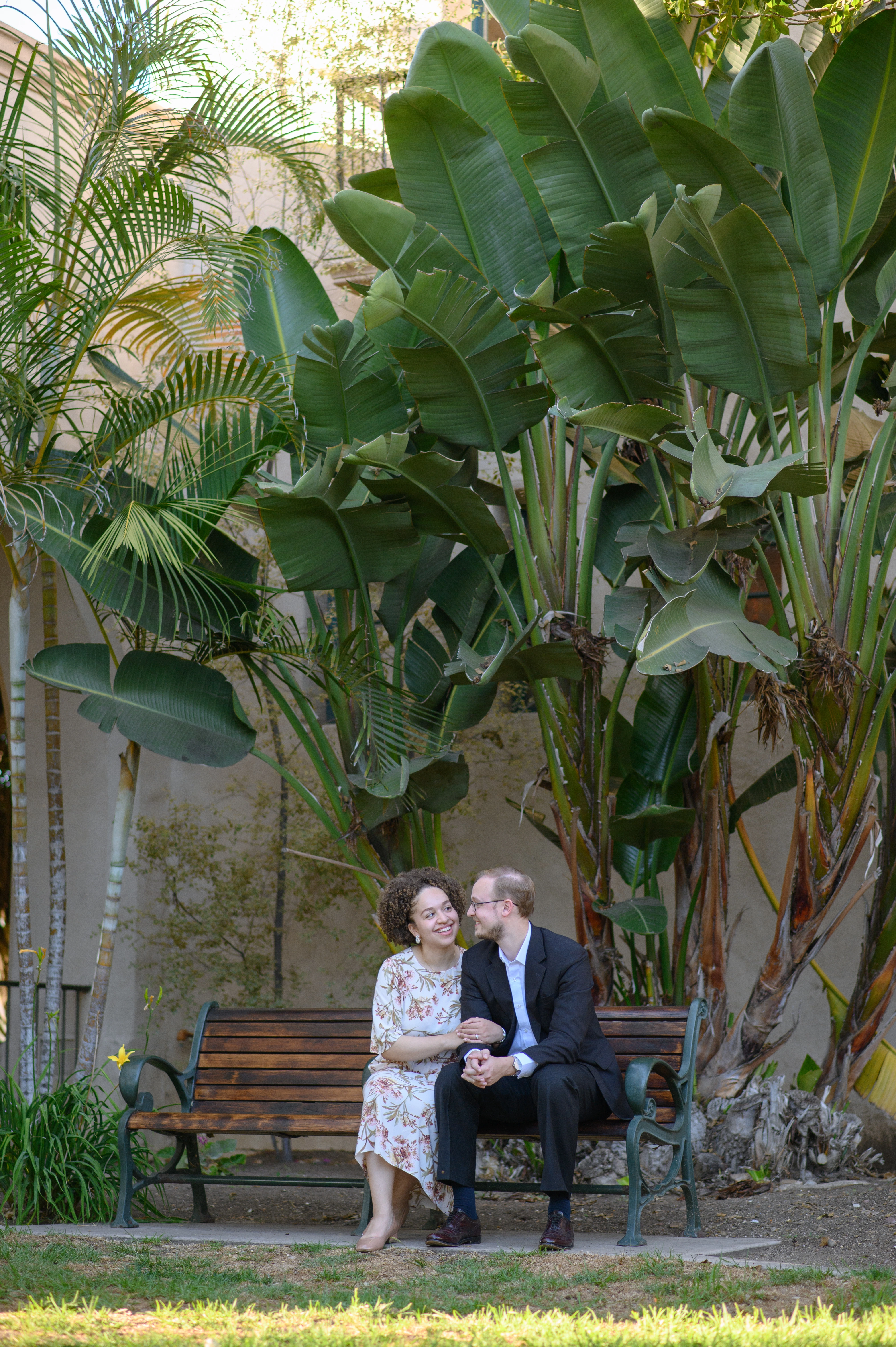 A couple sits closely together on a bench surrounded by lush tropical plants in a serene outdoor setting