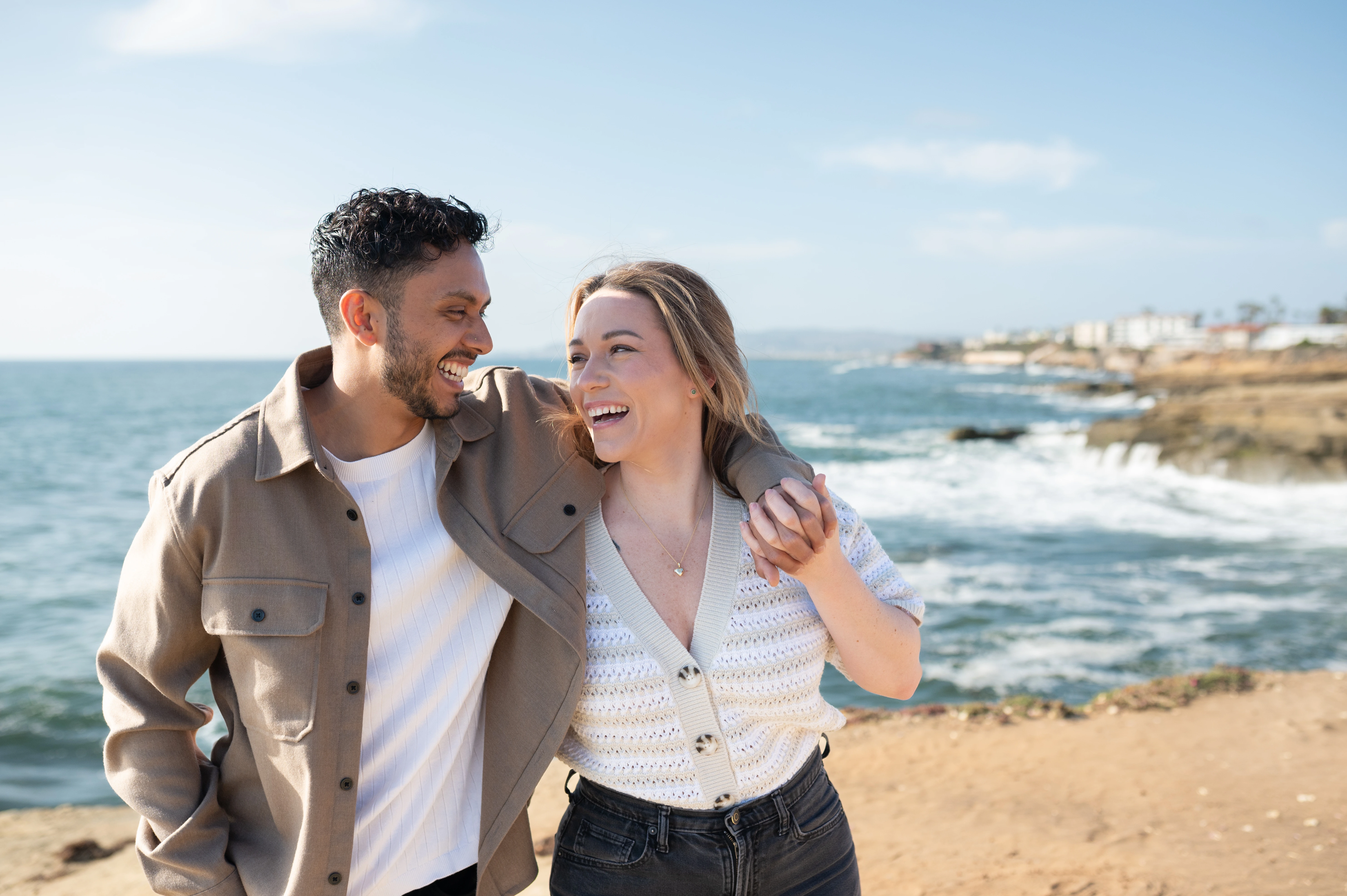 A joyful couple shares a candid moment during their engagement session at Sunset Cliffs in San Diego. The sun casts a warm glow on their faces as they walk hand in hand along the coastline, embodying love and happiness against the backdrop of the ocean waves. Perfect for showcasing the beauty of engagement photography.